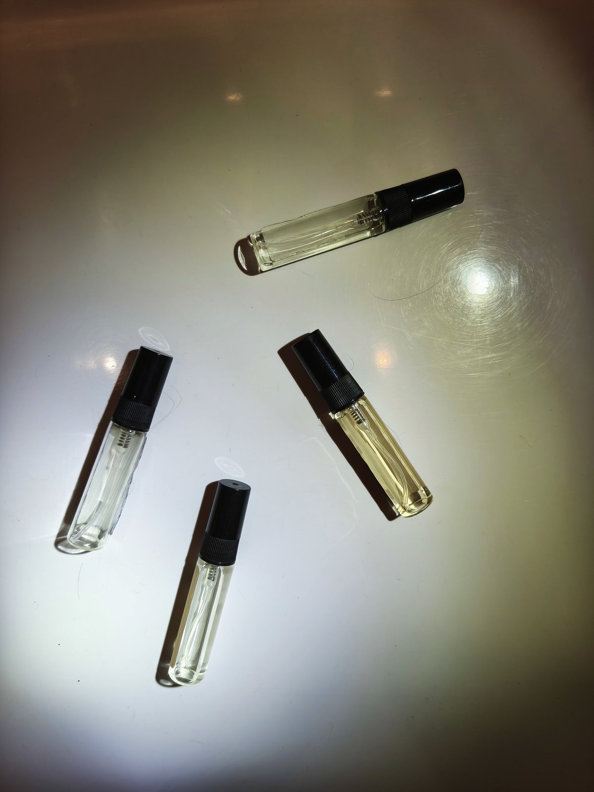Transparent Perfume samples with black caps on shiny surface under light. Close-up of cosmetic product with reflections, shadows. Glass fragrance bottle. Four vials, tester bottles. Vertical photo